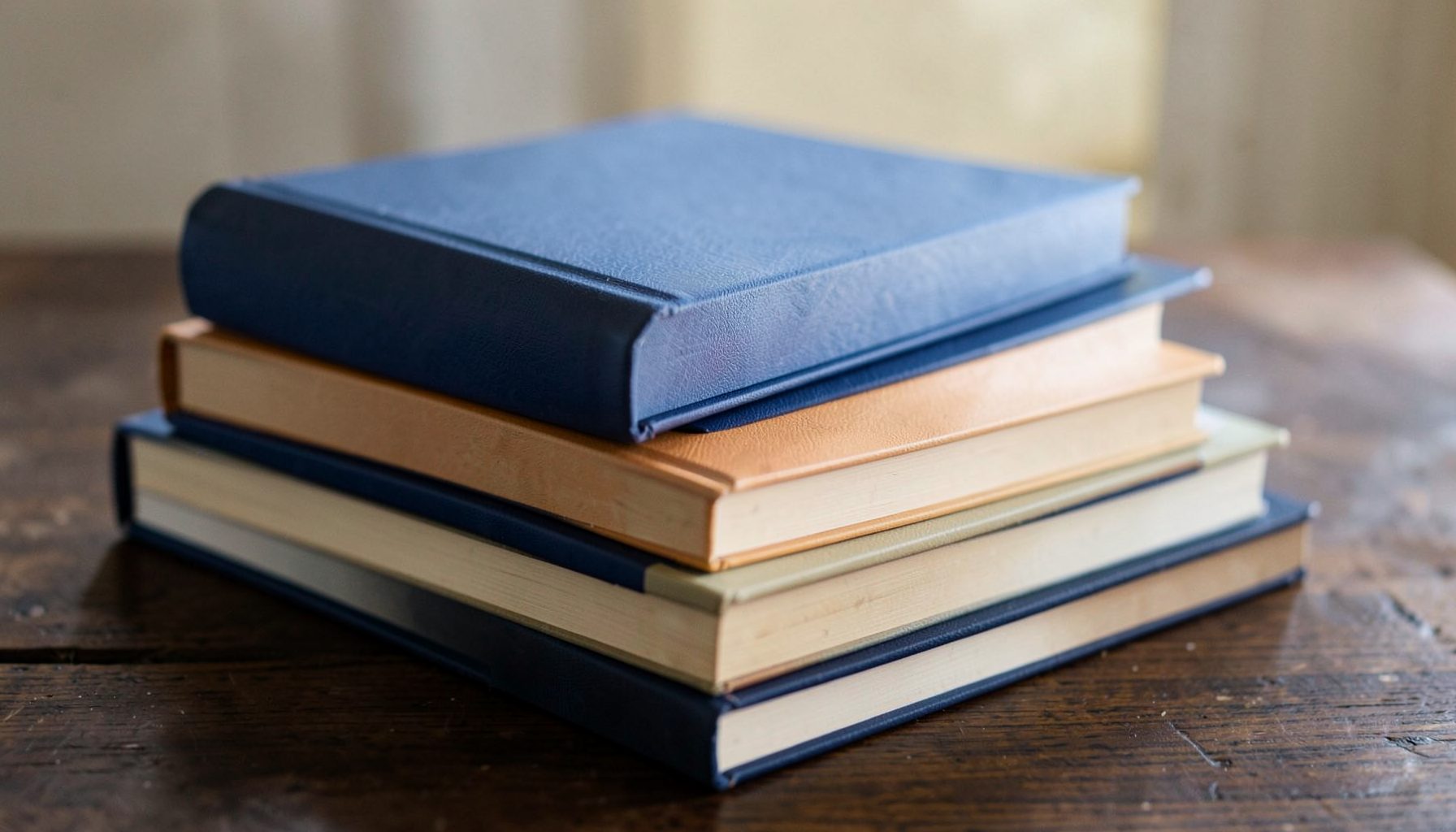 A stack of freshly printed hardcover books with crisp dust jackets in deep blues, warm ochres, and cream tones, resting on a dark surface